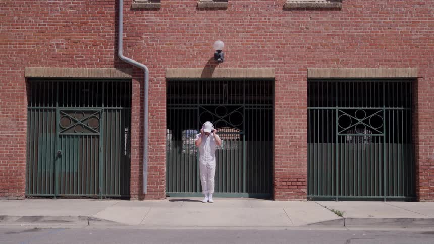 A man in an allwhite outfit dances and walks in front on a brick building radiating playful energy