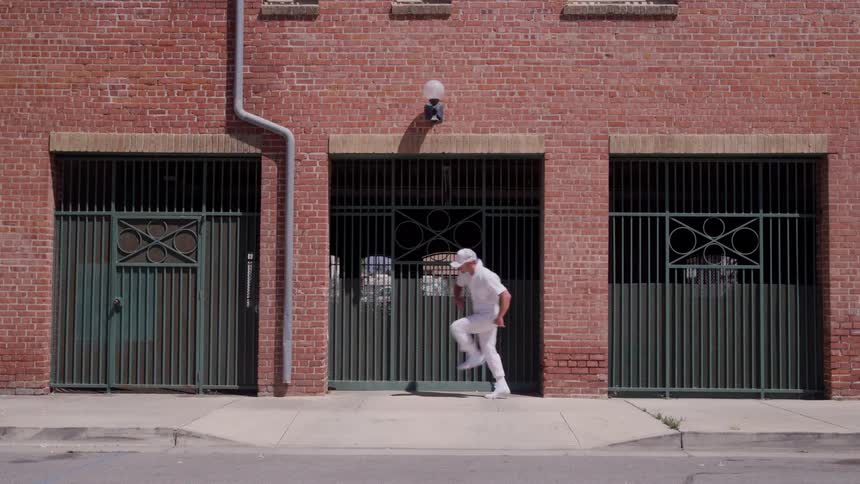 Man dressed in white walks while dancing in front of a brick building under the sun