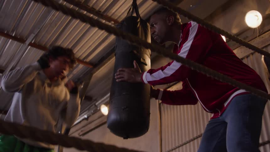 A young male boxer training with a punching bag held by a trainer in a boxing ring indoors during daytime