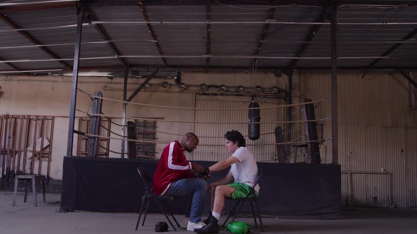 An adult man in a red track jacket and a younger man in green shorts sit facing each other on folding chairs in the foreground of a rustic boxing gym with the adult man attending to the younger mans hands in front of a boxing ring and punching bags