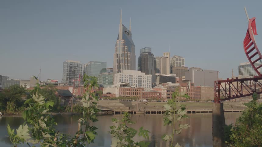 A city is shown across a calm river featuring the prominent tall skyscraper with the distinctive twin spires and several shorter buildings along the river bank with green foliage and a red metal structure visible in the foreground