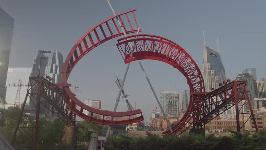 A large red metal sculpture composed of curved trusslike sections stands prominently in the foreground against a backdrop of a city skyline with tall modern skyscrapers under a clear sky