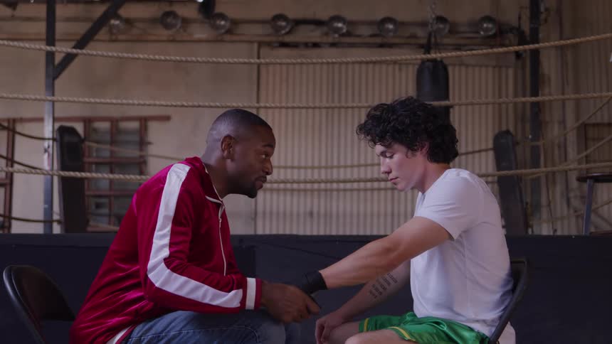 Two men sit on a bench beside a boxing ring in a gym at midday engaged in conversation as the black man wraps the wrist of the young white man preparing him for training