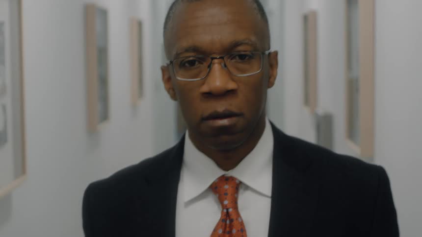 The video begins with a man in a business suit and glasses looking directly at the camera while standing in an office hallwayhe then looks down and the camera follows as he reaches down and picks up a box containing frames and other items
