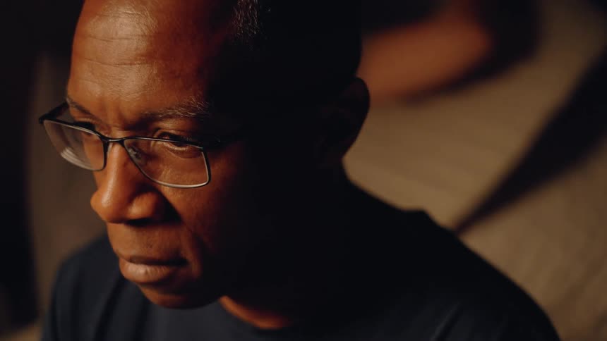 The video is an extreme closeup of a mans face heavily shadowed on one side highlighting his serious expression and glasseshe is looking down or offcamera conveying a mood of deep thought or concern in a dimly lit setting