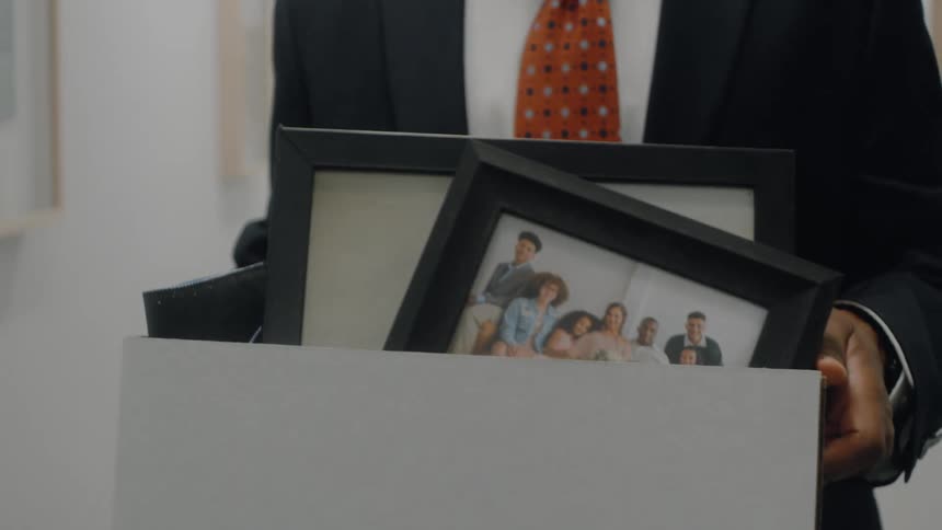 The video clip shows a closeup of a person in a suit and an orange patterned tie carrying a boxinside the box are personal items including a framed photograph of a group of people suggesting they are leaving a job or moving out of an office