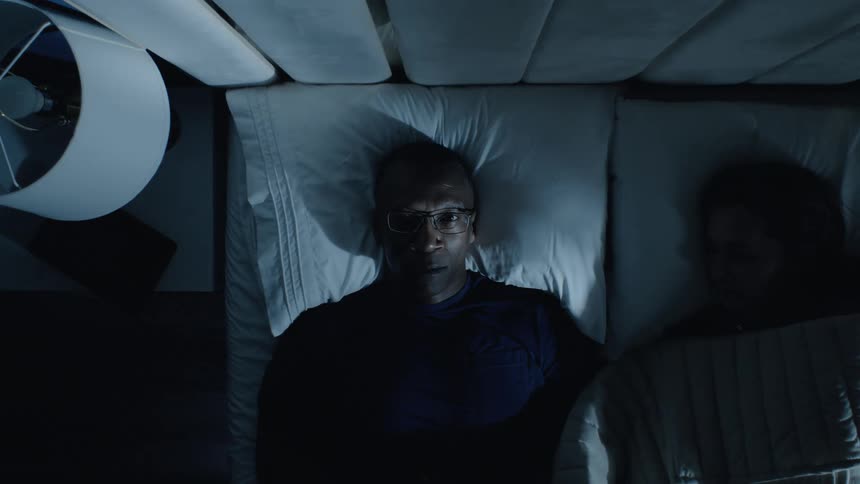 Man lies awake from insomnia while partner sleeps beside in a bedroom