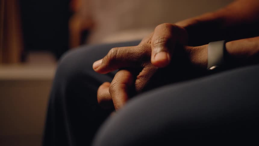 The video is a closeup shot focusing on a persons hands resting on their lapthe person is wearing dark clothing and a black wristband or watch and their fingers are interlaced or clasped conveying a sense of tension or deep thought