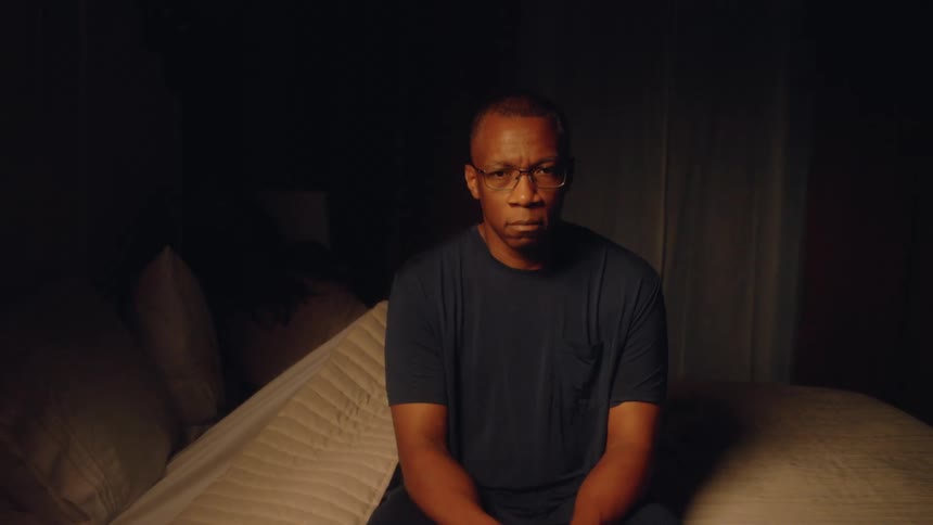The video clip shows a man sitting on the edge of a bed in a dark room looking directly at the camera with a somber expressiona woman is briefly visible beside him touching his shoulder and speaking before she lies down suggesting a moment of quiet