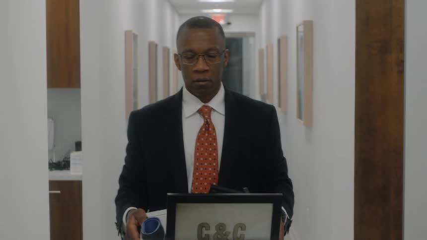 The video features a man in a business suit and glasses walking down a long white office hallway carrying a box of personal belongingshe maintains a sober serious expression while looking straight ahead suggesting he may be leaving his job