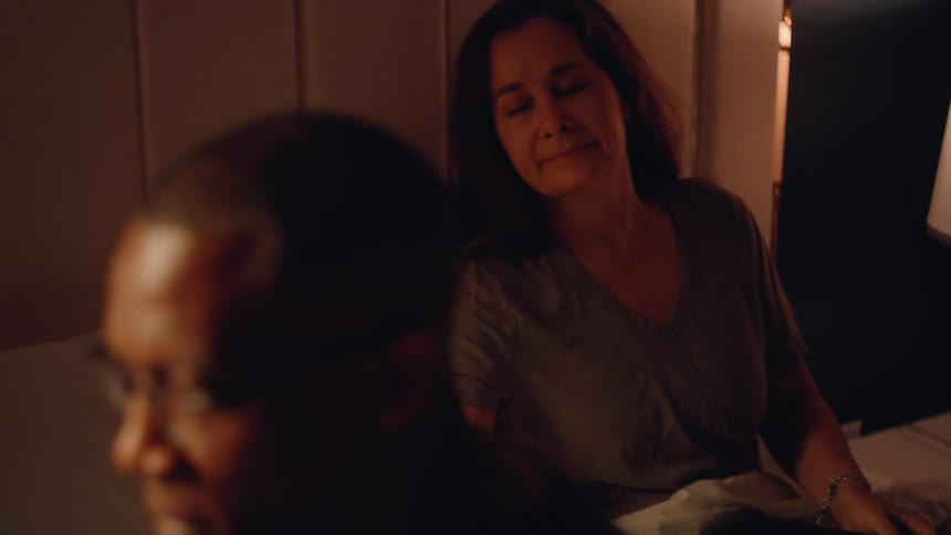 A middleaged couple spends a quiet night together in their bedroom the woman sits comfortably on the bed reading a book then turns to talk to the man seated at the bedside wearing reading glasses and listening attentively