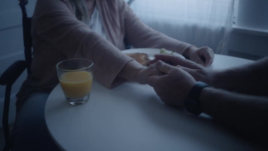 An elderly woman sits at a dining table indoors enjoying a sandwich and a cup of juice she smiles warmly as someone gently taps her arm sharing a lighthearted moment together