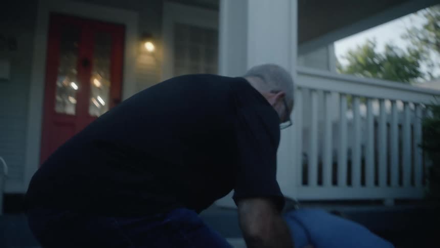 A bald whitehaired man in a black shirt quickly kneels to embrace and assist a woman in a blue shirt who has fallen on the steps of a house with a red front door