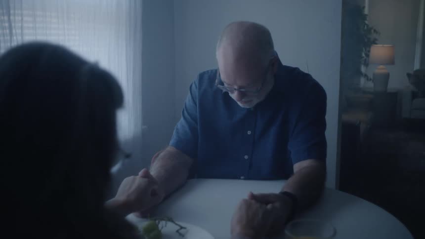 The video clip shows a man bringing a woman who is already seated at a small table for breakfast a glass of juice and a pillafter the woman accepts the medication the man sits down opposite her and they clasp hands across the table sharing a momen