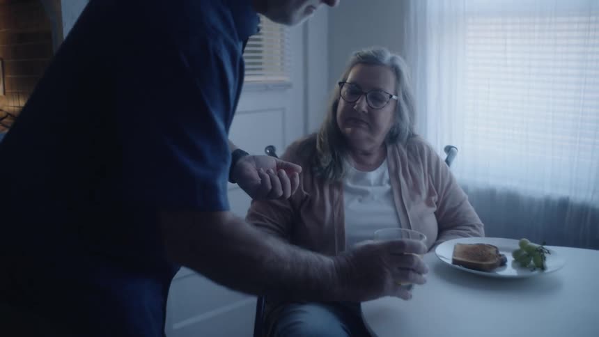 The video shows an older man preparing medication in the kitchen before walking to the dining areahe gives the pill to an older woman who is seated at the table with breakfast and then they hold hands across the table in a comforting gesture