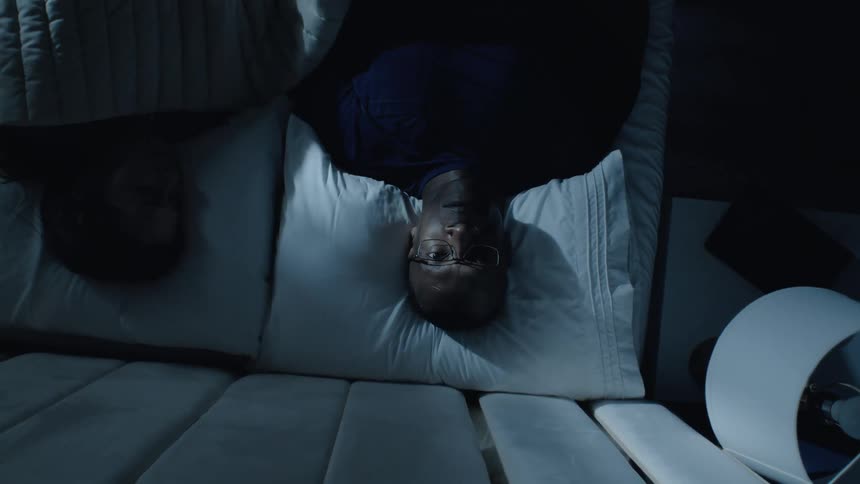 The video is an overhead shot of a man lying awake in bed in a dark room next to a sleeping personhe is lying on his back with his eyes open looking up at the ceiling with a troubled expression suggesting insomnia or deep distress