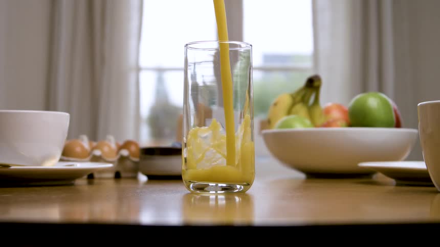 A stream of orange liquid is poured from above into a tall clear glass positioned on a wooden table which is also set with a bowl of fruit a tray of eggs and white beverage cups in front of a bright window