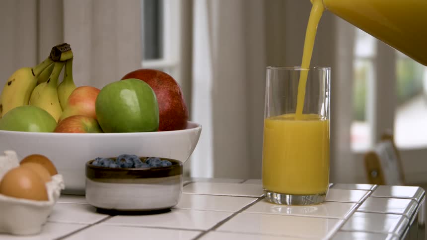 Closeup of orange juice poured into a glass next to a bowl of fresh fruit