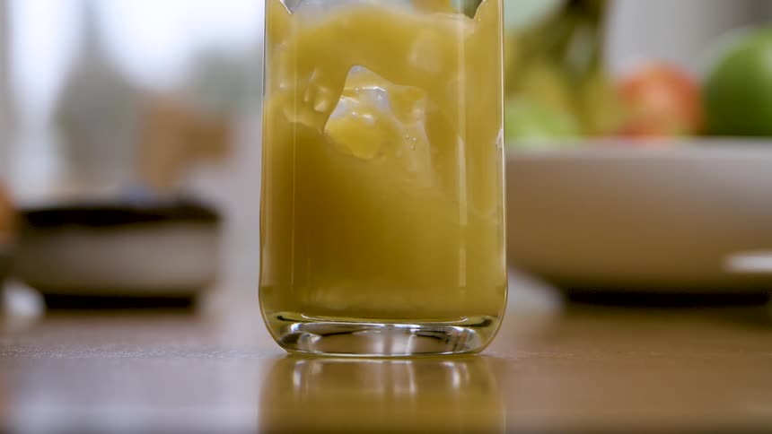 A golden smoothie or juice swirling as its poured into a clear glass on a wooden table with a blurred fruit bowl in the background