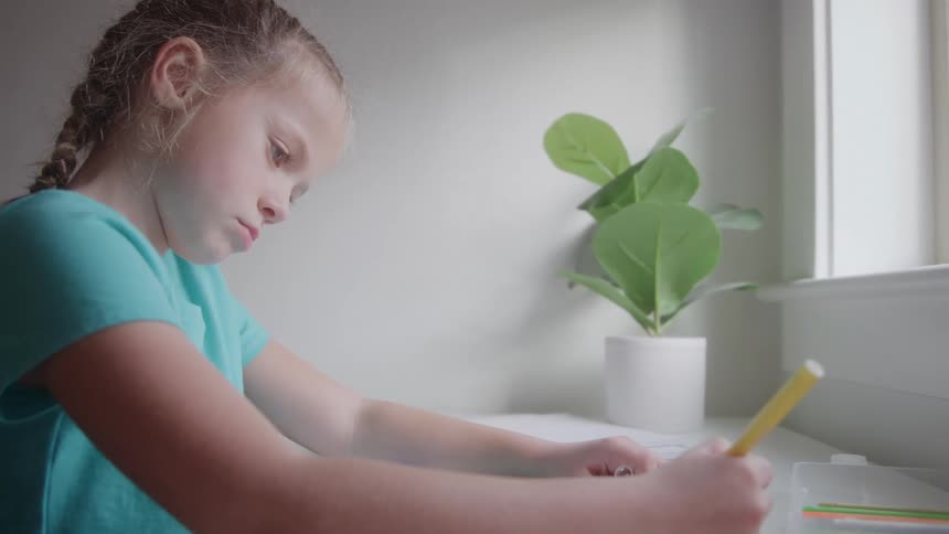 Young girl in a teal shirt sits by a bright window drawing with a yellow marker her expression focused and thoughtful with a potted plant in the background