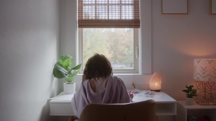 Person sitting with their back turned on a brown chair writes at a white desk near the window surrounded by soft light and plants