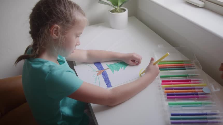 A young woman in a teal tshirt with braids sits at a white desk and colors a drawing of a tree with a yellow marker with an open case of colorful markers lying next to her