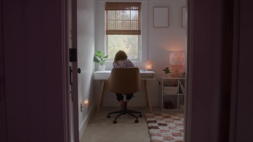 A woman with curly hair sits facing a window at a desk with a laptop and lamp in a room with a patterned rug with the scene framed by a dark doorway and illuminated by the bright window light