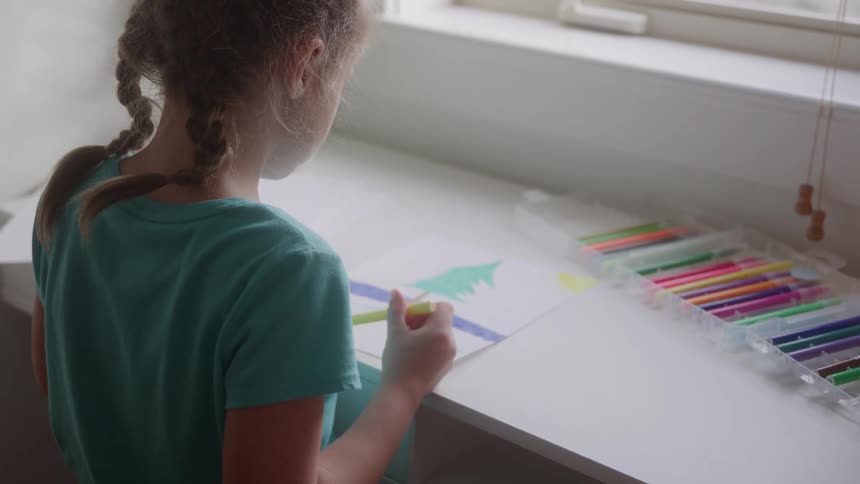 A little girl draws a tree on paper at a table by a window