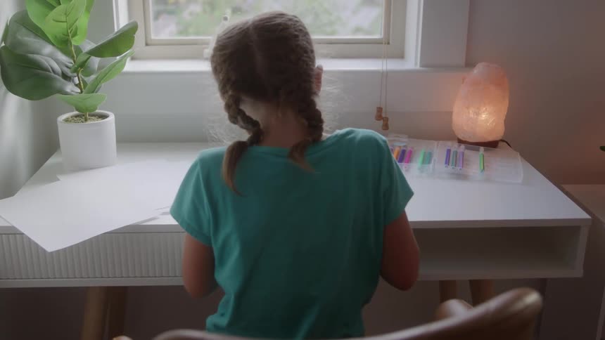 A young woman with braided brown hair and a teal tshirt sits at a white desk and draws near a window with a pink himalayan salt lamp and potted plants decorating the space