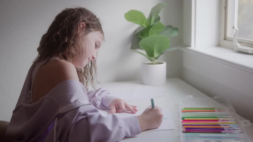 A young woman with long wetlooking hair and a light purple offshoulder sweatshirt sits at a white desk and draws on paper with a green colored pencil next to an open marker set and a potted plant