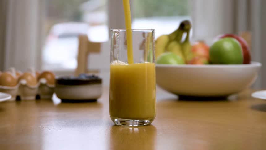 Closeup of orange juice being poured with fruit and eggs in the background