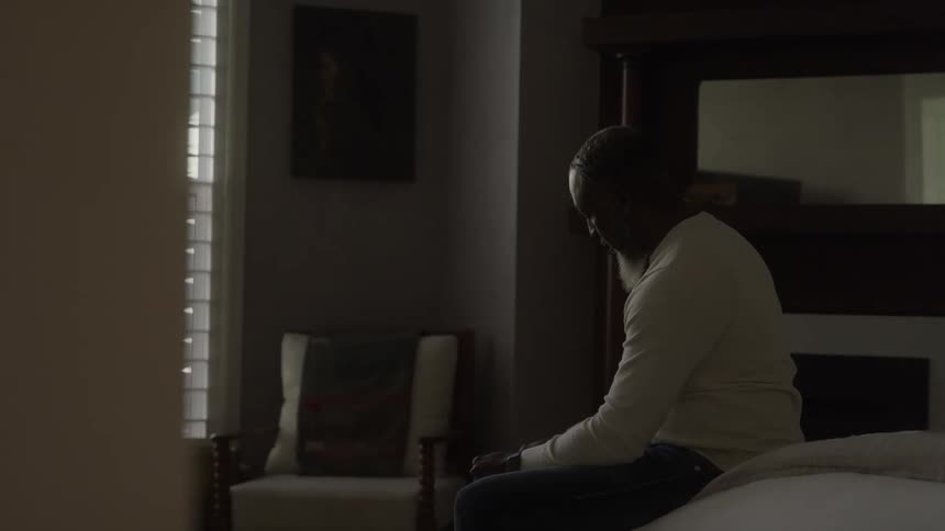 Man sitting on the edge of a bed in a softly lit room looking thoughtfully to the side