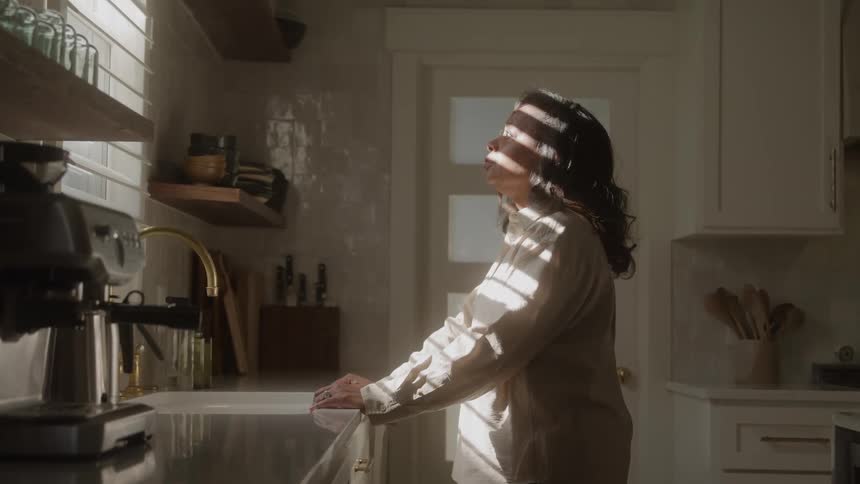 A depressed woman stands by a sunlit kitchen counter