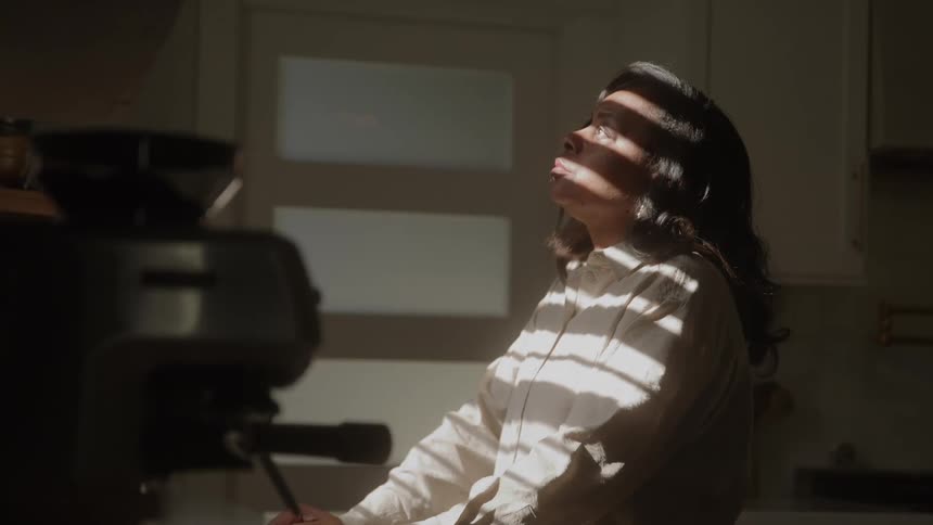 A woman stands in striped window light at a kitchen counter looking thoughtful and calm