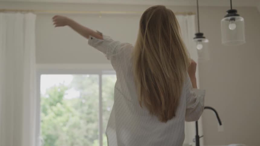 A woman with long hair twirls joyfully in a bright kitchen smiling with carefree energy