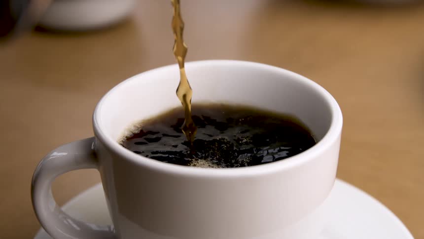 Liquid coffee is poured into a white ceramic mug resting on a matching saucer causing the liquid to ripple and foam until the pour stops and drops of coffee fall from above