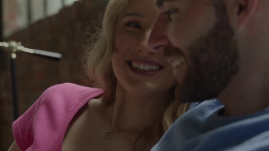 A smiling longhaired woman in a pink top and a bearded man in a blue shirt embrace and laugh together while sharing a moment in a brightly lit indoor space