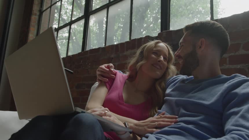 A man and a woman relax on a couch looking at a laptop while he holds a card both appearing content and engaged