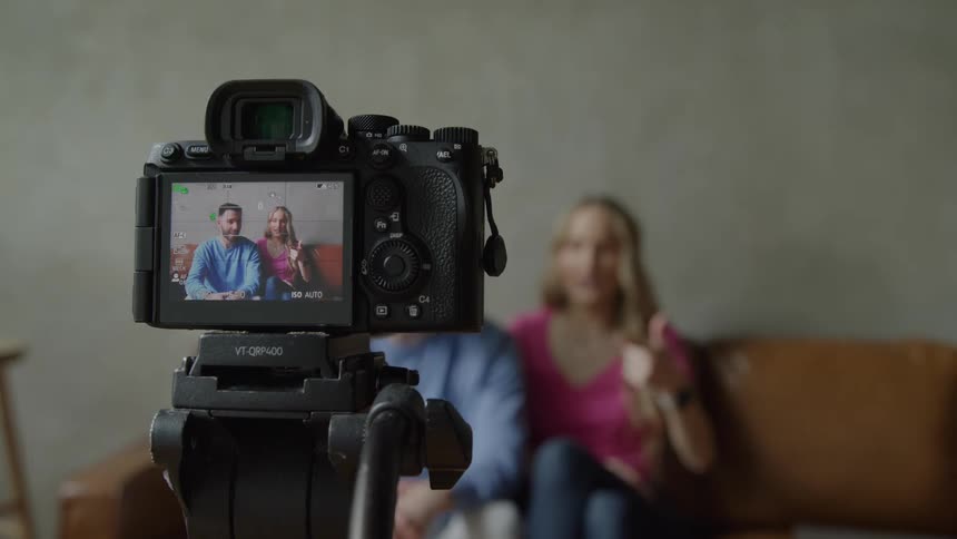 A camera on a tripod films a couple talking while sitting on a couch indoors