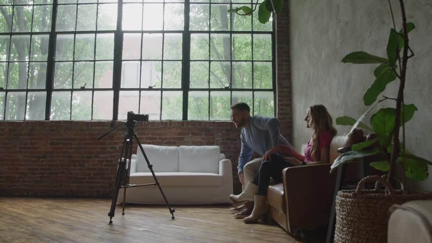 A man walks away from a camera mounted on a tripod and sits down next to a woman on a brown couch and they both smile and talk while facing a large industrial window in a loftstyle room with an exposed brick wall