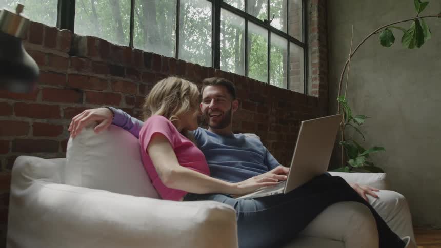 Couple shop online together on a laptop while sitting on a sofa at home