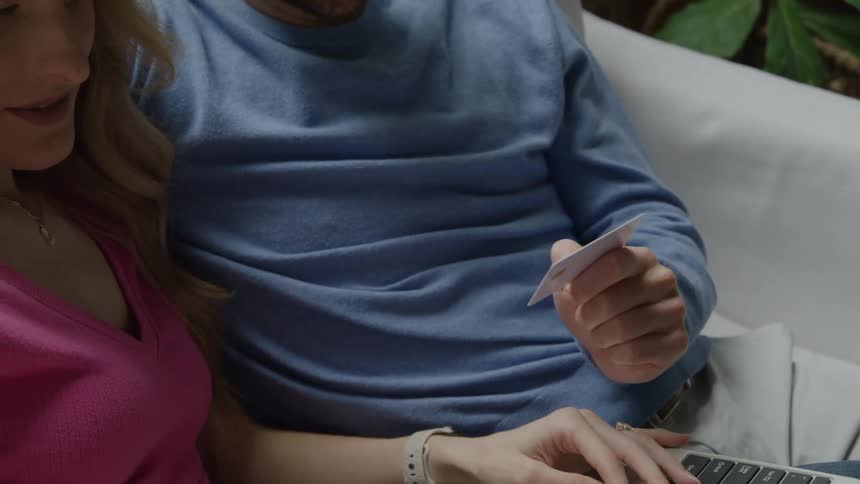 A young couple sits together on a couch smiling at each other as the woman makes an online purchase using a laptop while the man holds an atm card in his hand