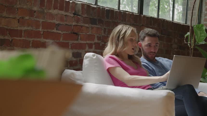 A man and a woman sit together on a white couch in front of a brick wall and a large window with the man holding a laptop and the couple discussing something on the screen while pointing and gesturing around the room