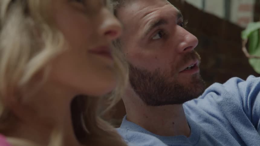 The video clip features a closeup of a young couple sitting together possibly on a couch with plants in the backgroundthe man and woman are looking and talking to each other and then smiling warmly indicating a happy or engaged conversation