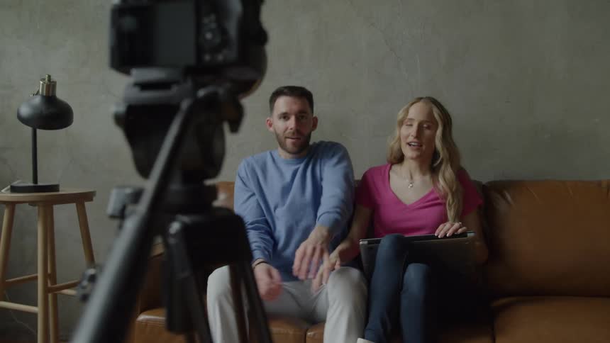 A man sets up a camera on a tripod while a woman in a pink top sits on a leather couch smiling until he joins her for a picture