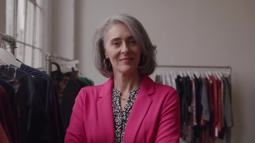 A woman with gray hair and a pink blazer stands in the middle of a retail space with clothing racks visible on both sides and looks directly at the camera with a warm smile