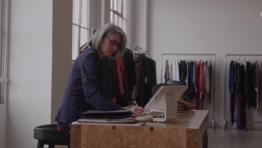 Grayhaired woman taking notes at a store counter with hanging clothes in the background
