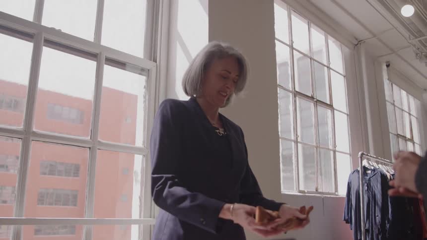 Grayhaired woman in a navy blazer smiles as a customer hands her an item from across a counter beside tall industrial windows