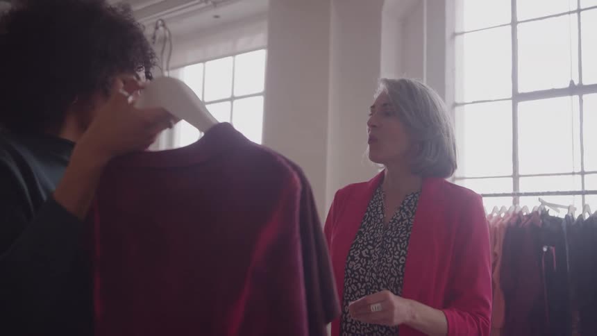 A woman in a pink blazer gestures and talks to a second woman who holds up a dark red shirt or sweater on a white hanger and looks at the garment while standing in a bright room with large windows in the background