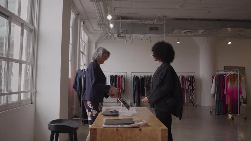 A woman walks through a store and approaches the cashier ready to make payment for her shopping with a relaxed smile and a few items in hand